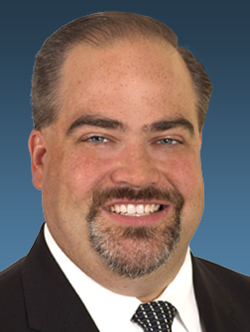 headshot of man with goatee in black suit jacket with white shirt and black tie